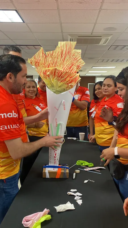 Equipo con playeras naranjas sosteniendo una antorcha de papel que construyeron juntos, entre sonrisas